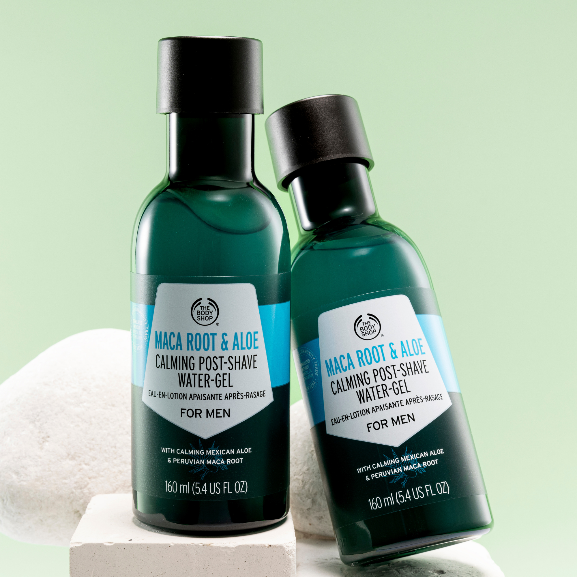 Two dark green bottles of The Body Shop Maca Root & Aloe Calming Post-Shave Water-Gel, one standing upright and the other tilted, on a light green background with a white towel and stone prop.