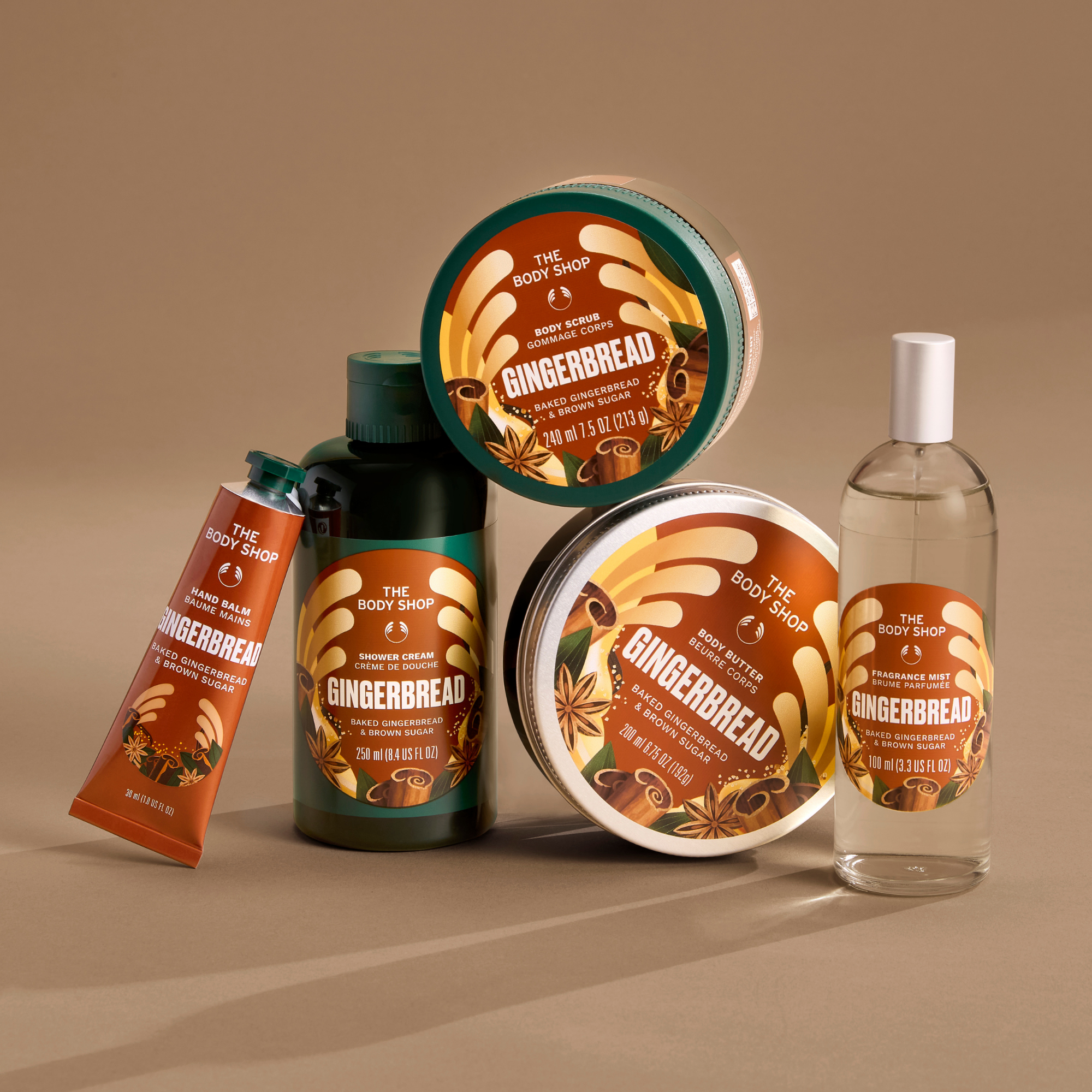 A set of The Body Shop Gingerbread body care products arranged on a warm beige studio background, including a tube of hand wash, a dark bottle, a clear bottle, and two round jars with green and orange labels.