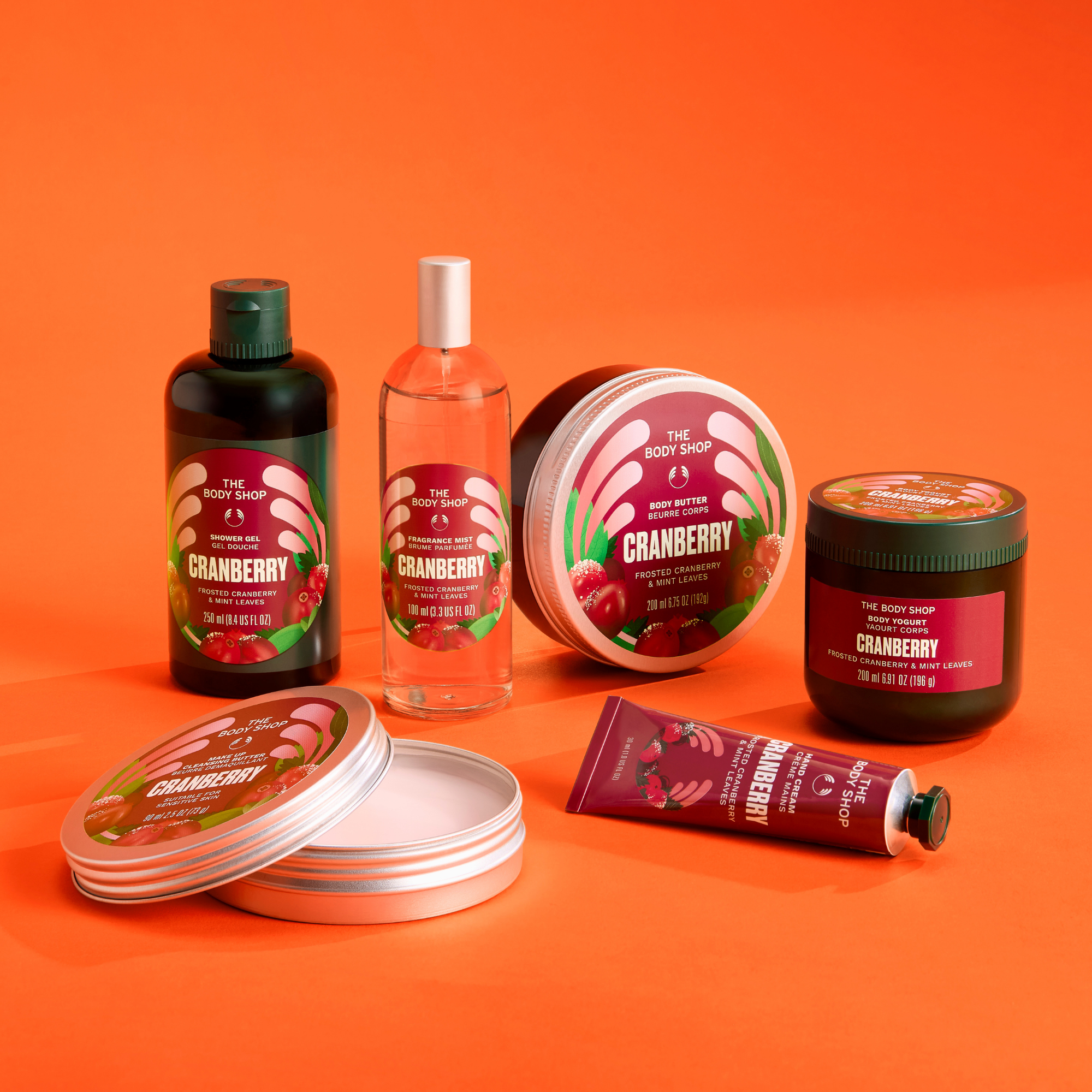 A set of The Happy Campfire bath and body products in a cranberry scent, including a dark bottle, a pink spray bottle, a round tin, a jar, and a tube, arranged on an orange background.