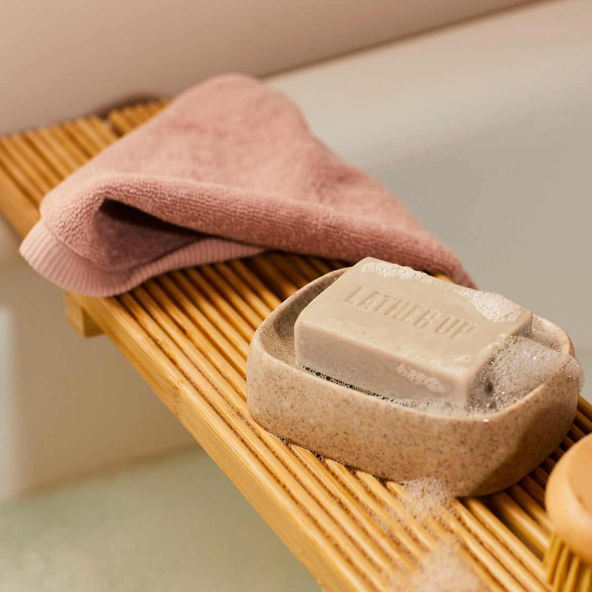 Bar of soap with suds in a wooden soap dish next to a pink towel on a wooden surface.