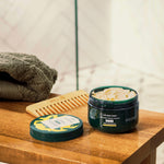 Jar of body scrub and lid on a wooden surface with a towel and comb in the background.
