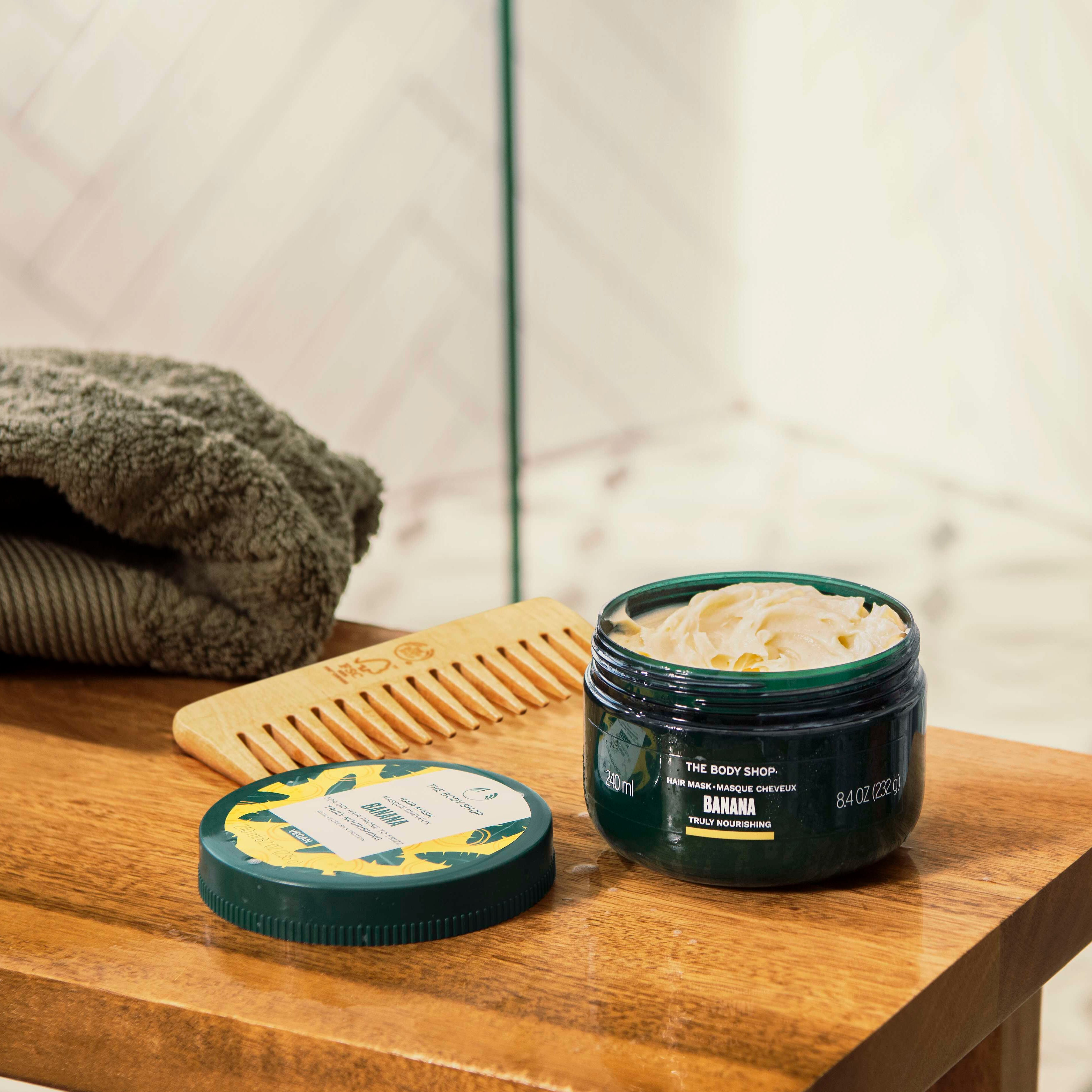 Jar of body scrub and lid on a wooden surface with a towel and comb in the background.