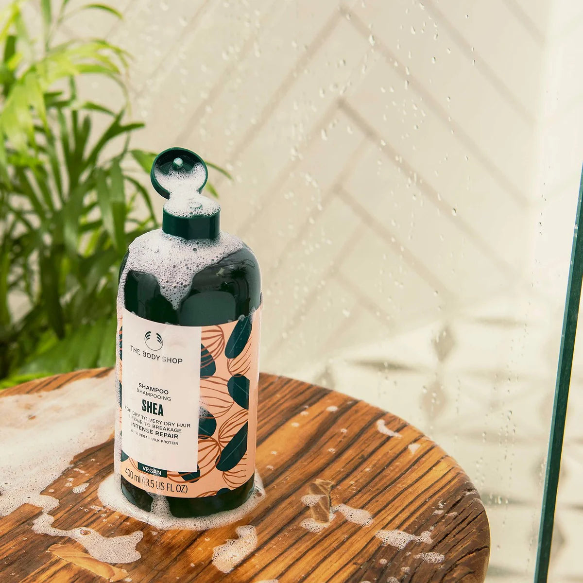 The Body Shop Shea Shampoo bottle filled with dark green liquid and topped with white foam, set on a wooden surface with soap residue inside a shower glass door.