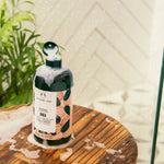 The Body Shop Shea Shampoo bottle filled with dark green liquid and topped with white foam, set on a wooden surface with soap residue inside a shower glass door.