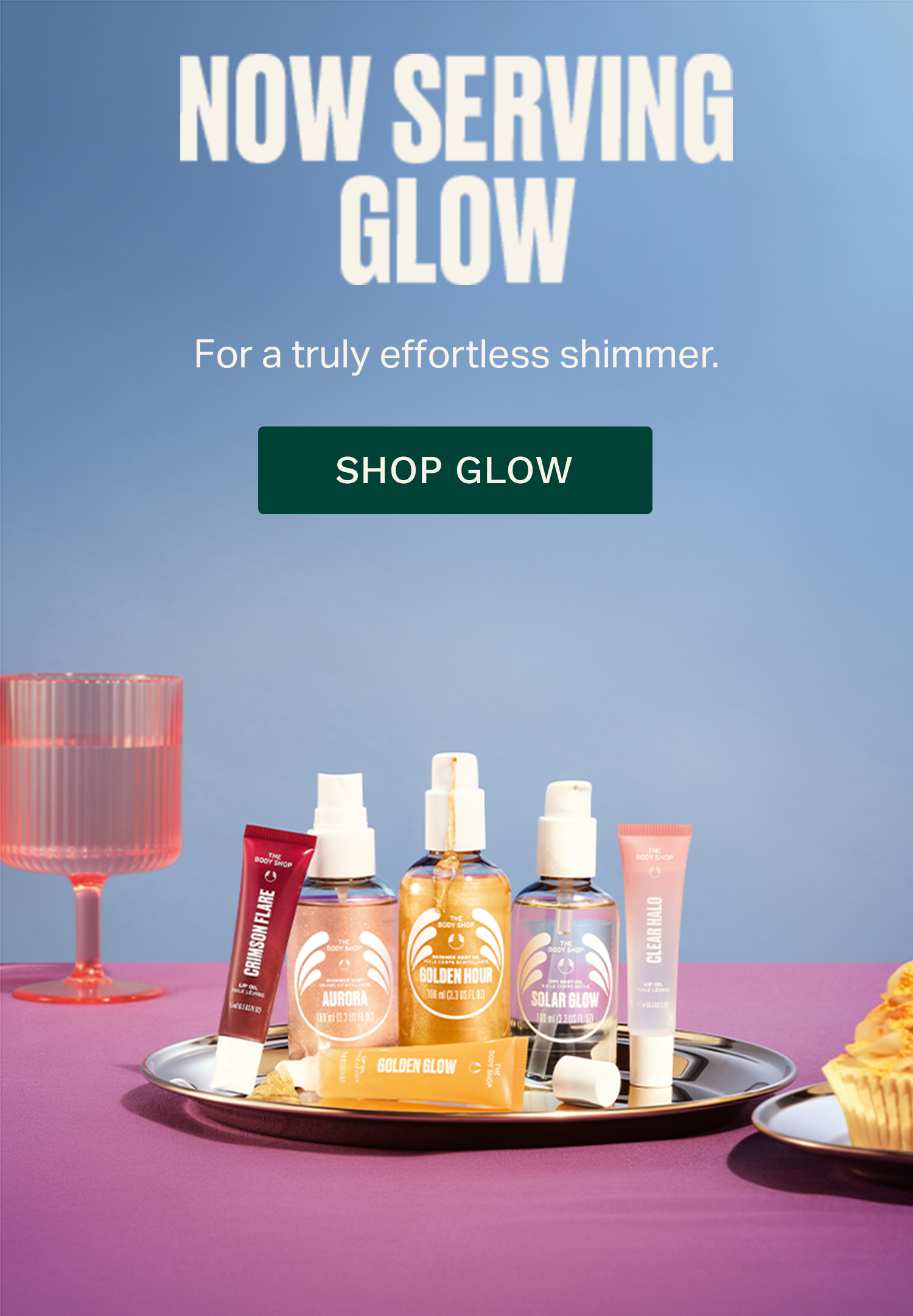 A promotional ad for GLOW cosmetics showing several product bottles and tubes arranged on a silver tray over a purple surface, with a pink glass and a cupcake in a studio setup. Large text at the top reads 'NOW SERVING GLOW' and 'SHOP GLOW'.