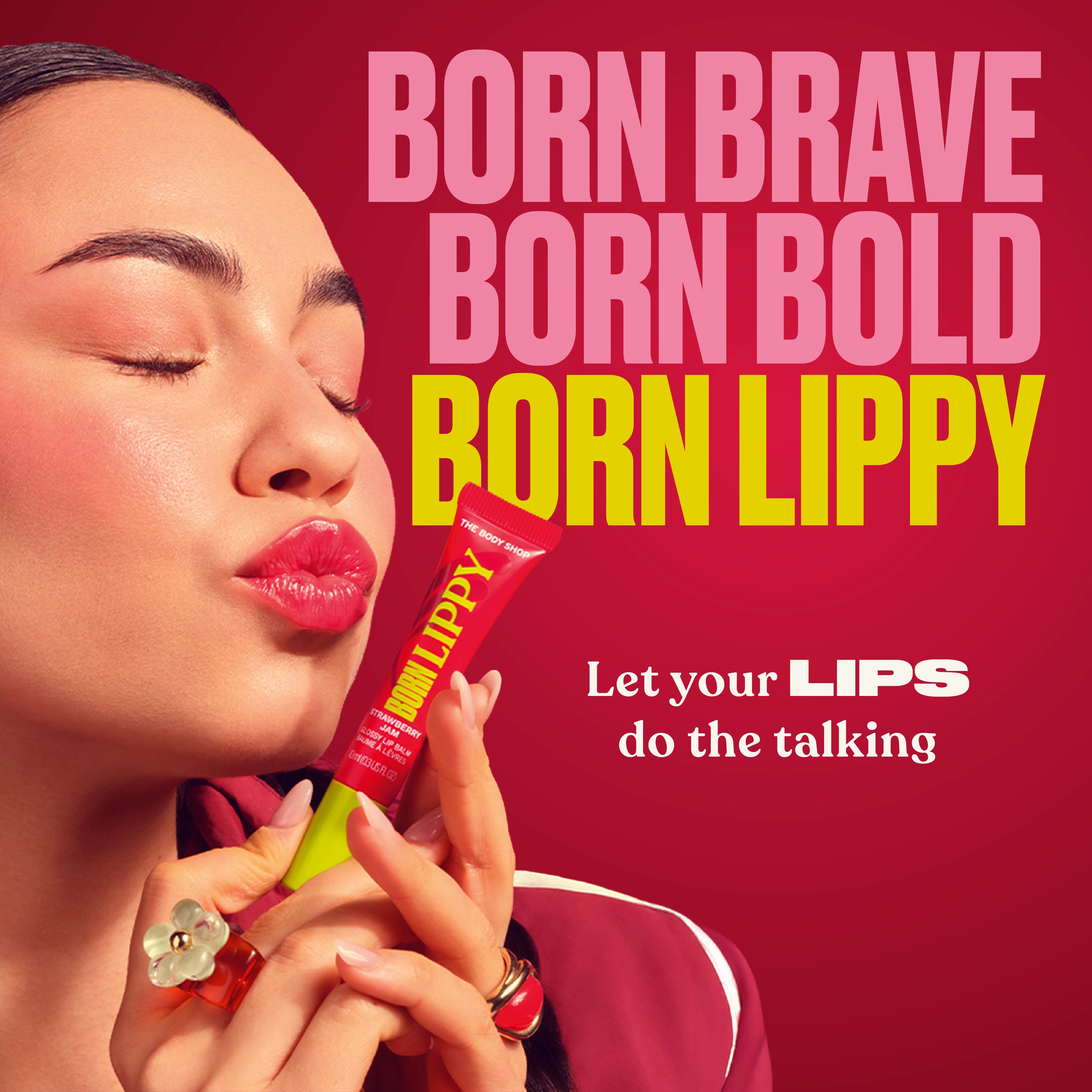A close-up promotional photo of a woman holding a red tube of lip product against a bright pink background with large text reading 'BORN BRAVE BORN BOLD BORN LIPPY' and smaller text 'Let your LIPS do the talking.' The product tube is visible and the brand/product line name appears on both the tube and the headline.