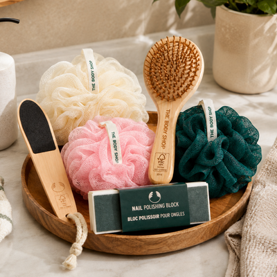 A bath and nail care gift set from The Body Shop arranged on a round wooden tray. The set includes a cream, pink, and dark green loofah, a wooden nail brush, a wooden nail file, and a green nail polishing block.