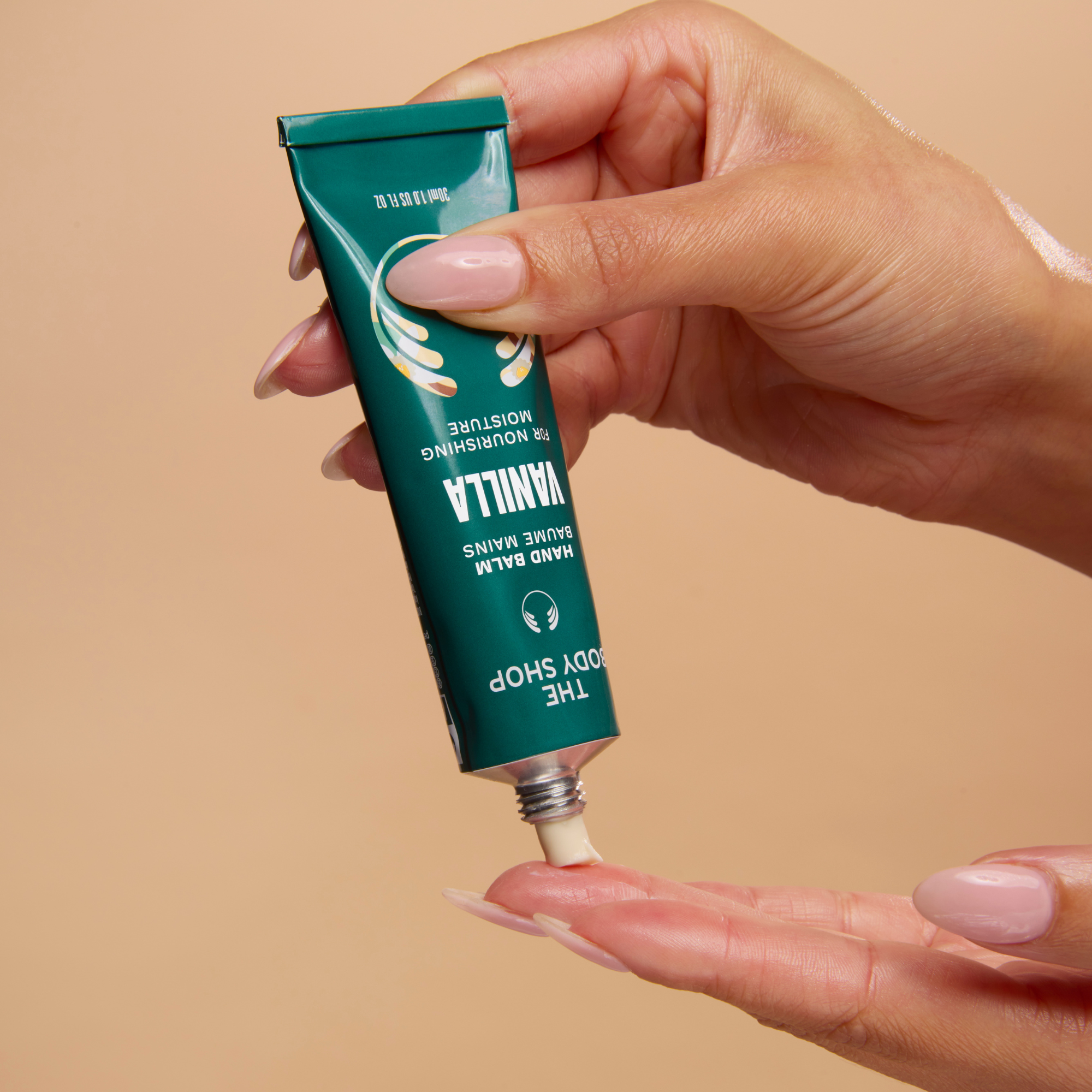 A hand holding a teal tube of The Body Shop Vanilla Hand Balm above another hand, showing the cream being squeezed onto a beige background.