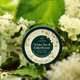 Body yogurt container labeled 'White Tea & Elderflower' by The Body Shop with green leaves and flowers in the background.