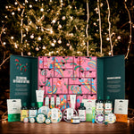 A festive gift advent calendar set displayed on a wooden table in front of a glowing Christmas tree. The set includes a large pink patterned calendar box with teal side panels, surrounded by assorted bath, body, and skincare products from F&F Beauty.