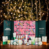 A festive gift advent calendar set displayed on a wooden table in front of a glowing Christmas tree. The set includes a large pink patterned calendar box with teal side panels, surrounded by assorted bath, body, and skincare products from F&F Beauty.