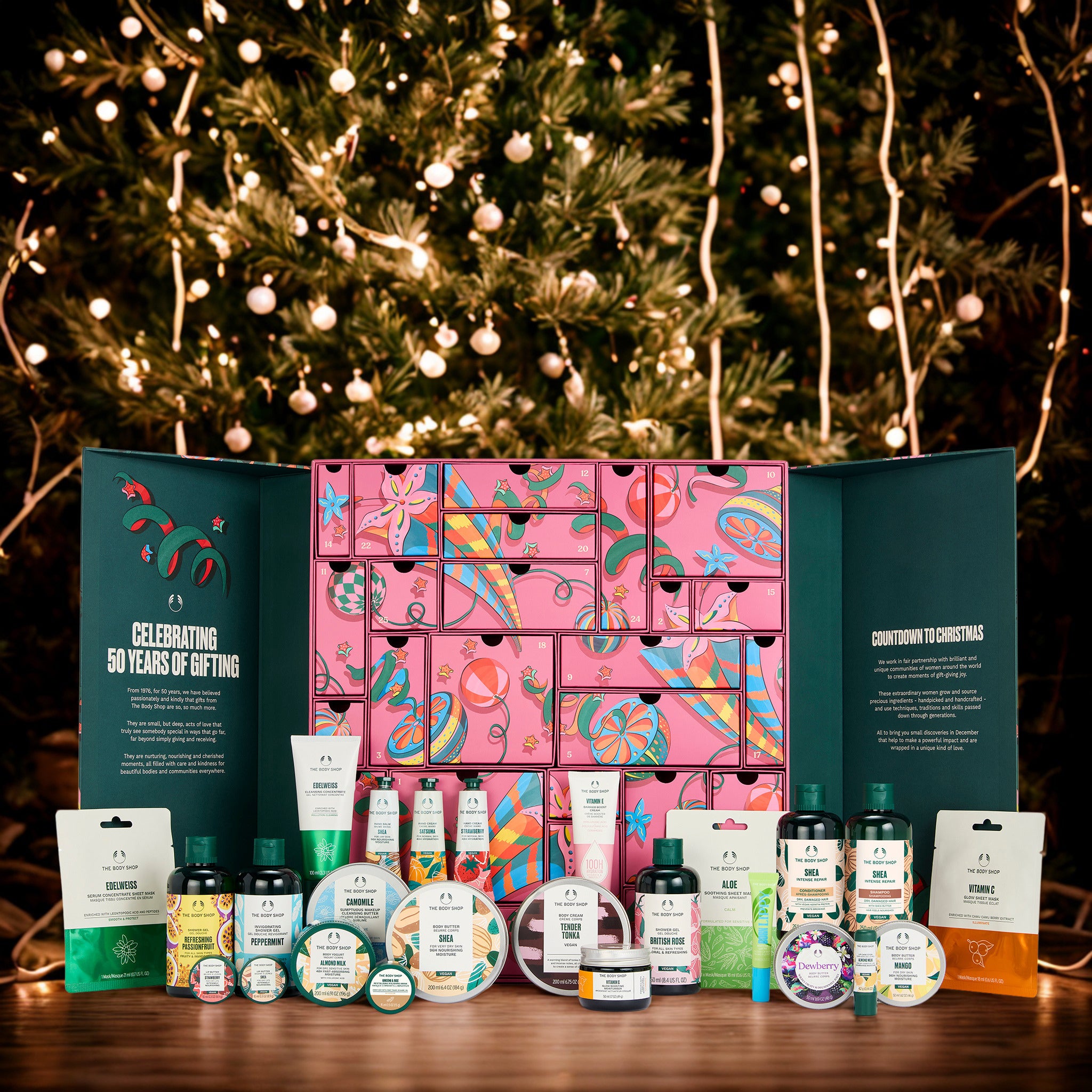 A festive gift advent calendar set displayed on a wooden table in front of a glowing Christmas tree. The set includes a large pink patterned calendar box with teal side panels, surrounded by assorted bath, body, and skincare products from F&F Beauty.