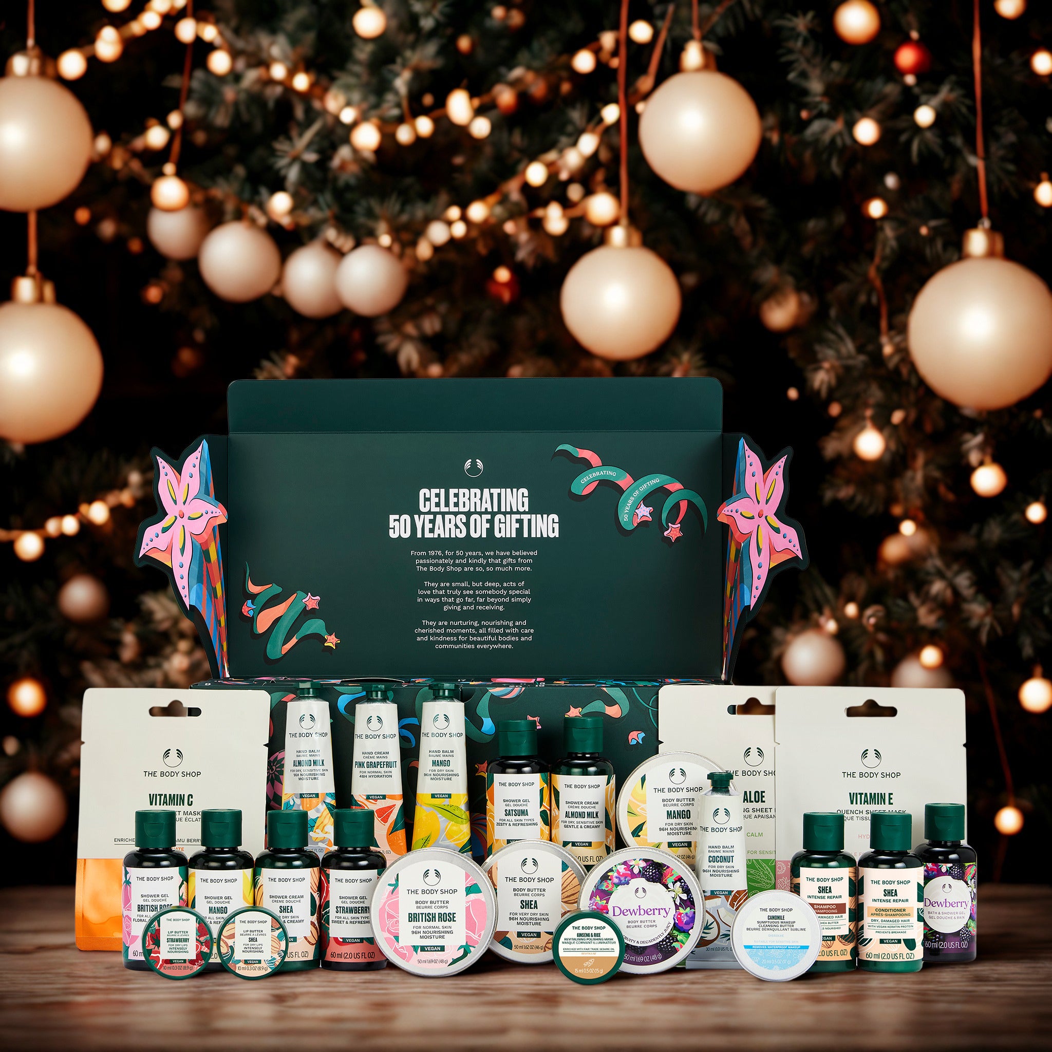 A curated gift set from The Body Shop displayed on a wooden surface in front of a decorated Christmas tree with warm lights and hanging ornaments. The set includes a large dark green gift box with floral artwork and the text 'Celebrating 50 Years of Gifting,' plus multiple bath, body, and skincare products such as lotions, creams, and bottles arranged in front.