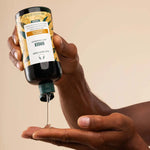 Hand holding a bottle of hair oil with visible brand label against a beige background