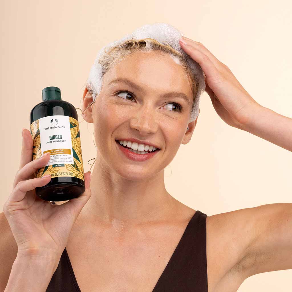 Woman with soapy hair holding a bottle of shampoo against a beige background