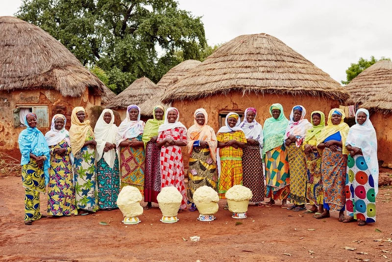 Group of women in colorful traditional attire standing in front of thatched-roof huts.